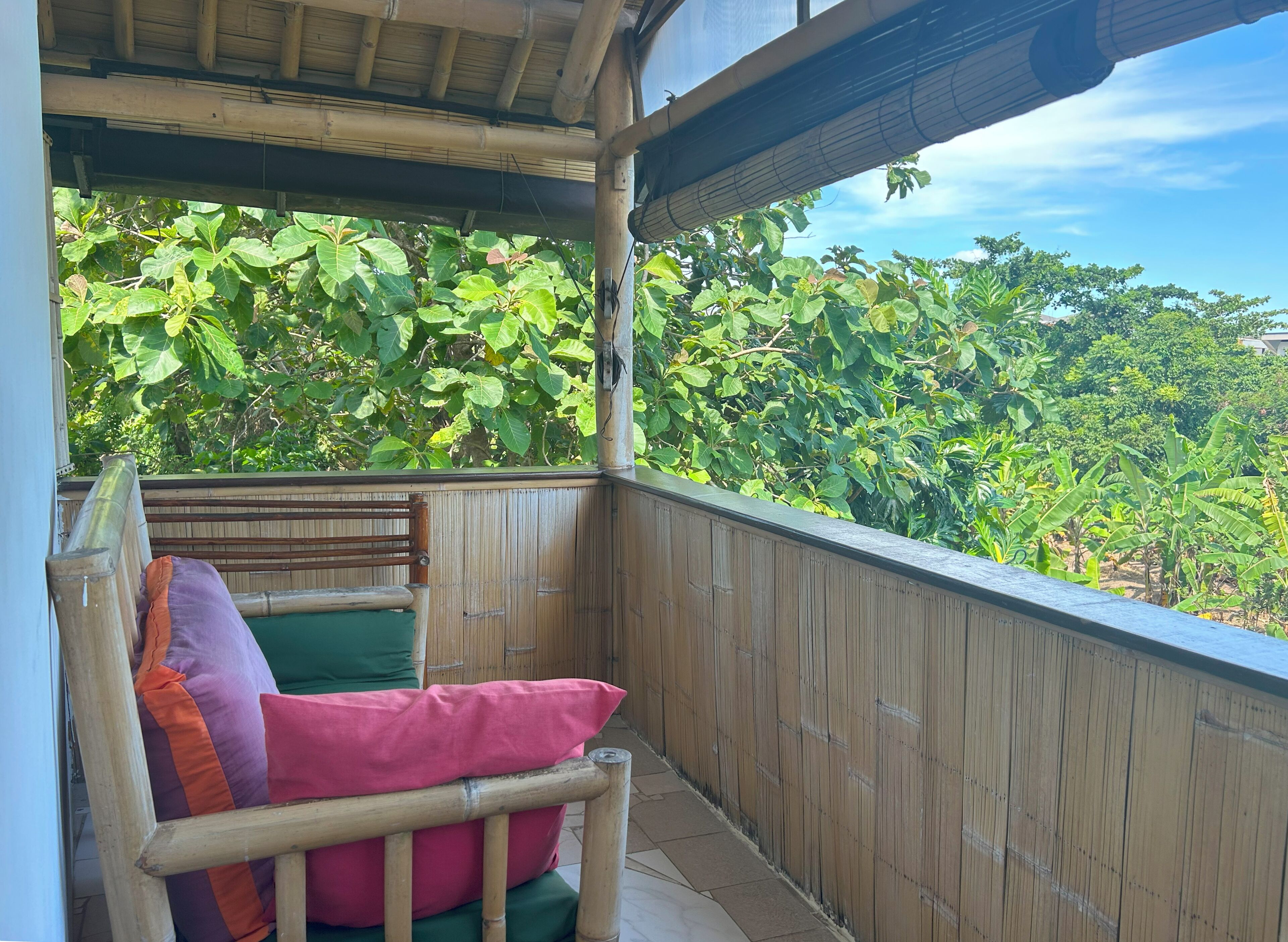 A wooden balcony with a cushioned bench and a view of lush greenery.