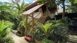 Eine hölzerne Treppe führt zu einem Haus mit einem Strohdach, umgeben von üppiger tropischer Vegetation.