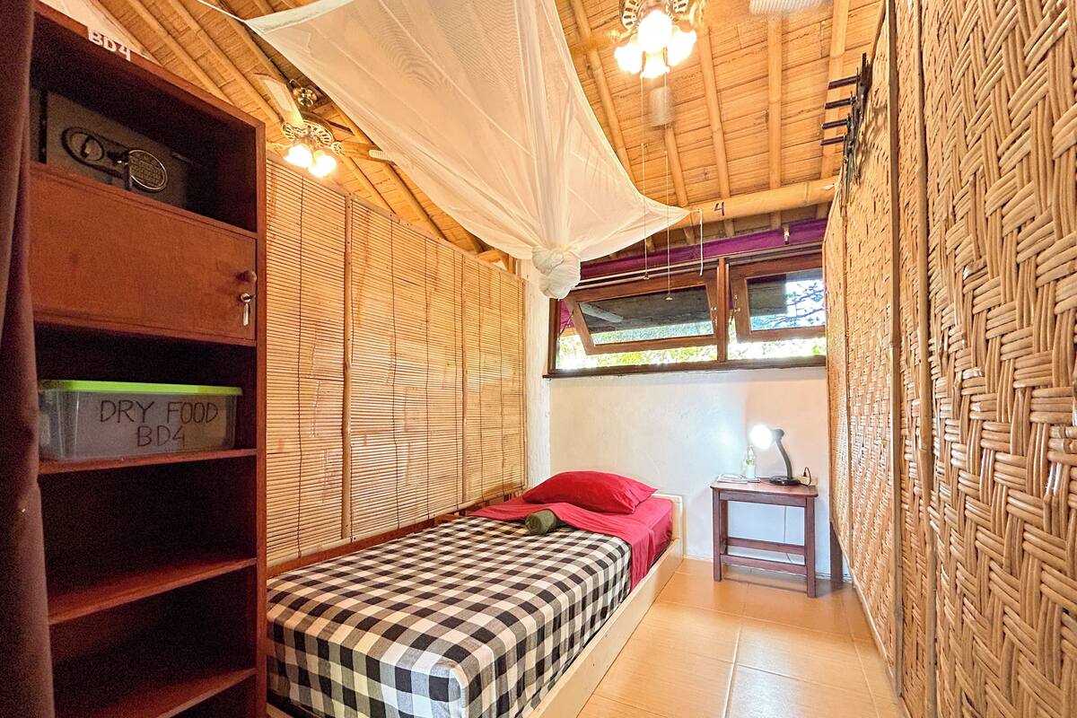 Una habitación pequeña con una cama, un estante con la etiqueta 'DRY FOOD', una mesita con una lámpara y una ventana con vistas.