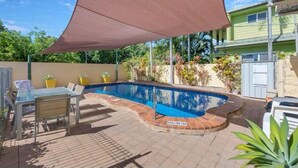 A swimming pool with a shaded area, a table with chairs, and potted plants.