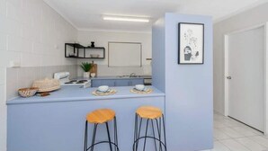 A compact kitchen with a blue countertop, two stools, a framed picture, and a white door.