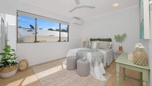 A bedroom with a large window, a ceiling fan, a bed with a gray blanket, and a small table with a plant.