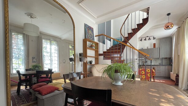 A spacious living area with a dining table, chairs, and a large mirror. A staircase leads to an upper level, and a kitchen is visible in the background. - Maison au Calme et au Vert au Cœur du Centre Ville (Vincennes)
