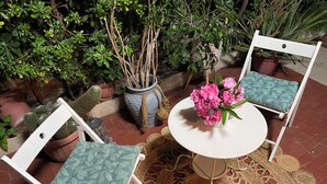A round table with a white top and a woven mat underneath, surrounded by two white chairs with green patterned cushions.