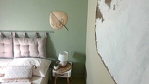 A bedroom with a bed, pillows, a bedside table with a lamp, and a wall with peeling paint.