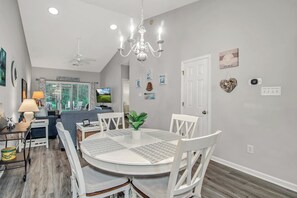 A dining area with a round table, white chairs, a chandelier, and a view of the living room.