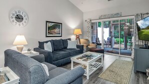 A living room with a sofa, coffee table, and a TV. There is a clock on the wall and a window with curtains. - TB 93C Wonderful 3/bed 2/bath Garden home in True Blue (Pawleys Island)