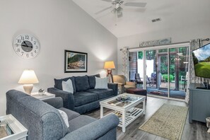 A living room with a sofa, coffee table, and a TV. There is a clock on the wall and a window with curtains.