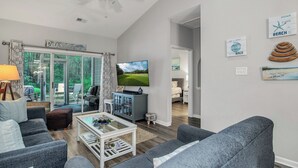 A modern living room with a flat-screen TV, grey sofas, a coffee table, and a ceiling fan. - TB 93C Wonderful 3/bed 2/bath Garden home in True Blue (Pawleys Island)
