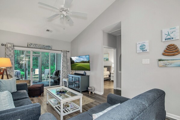 A modern living room with a flat-screen TV, grey sofas, a coffee table, and a ceiling fan.