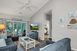 A modern living room with a flat-screen TV, grey sofas, a coffee table, and a ceiling fan.
