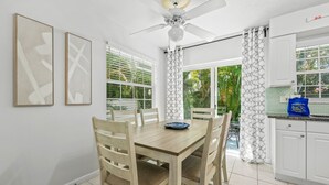 A dining area with a wooden table and chairs, a ceiling fan, and a kitchen with white cabinets and a tiled backsplash.
