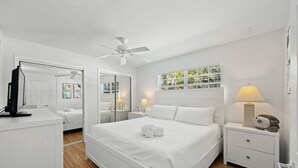 A white-themed bedroom with a bed, bedside tables, a ceiling fan, and a window with blinds.