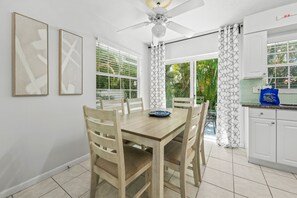 Een eethoek met een houten tafel en stoelen, een plafondventilator, en een keuken met witte kastjes en een betegelde spatwand.