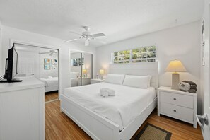 Een slaapkamer in witte tinten, met een bed, nachtkastjes, een plafondventilator en een raam met jaloezieën.