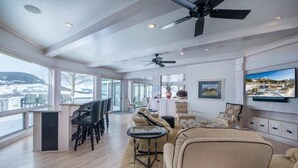 A spacious living area with a large window overlooking a snowy landscape, a ceiling fan, and a television mounted on the wall. - Spacious Mountain Manor with 9 bedrooms, 11 baths and Hot Tub (Crested Butte)