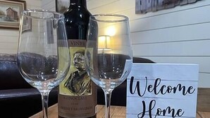 A bottle of wine and two glasses on a wooden table with a 'Welcome Home' sign. - PineyWoods House Forest Retreat (Bastrop County)