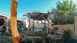 Eine Pergola aus Holz in einem Garten mit verschiedenen Pflanzen und einem Pool im Hintergrund.