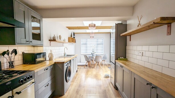 A modern kitchen with a built-in oven, microwave, and washing machine. There is a dining area with a table and chairs, and a window with blinds.