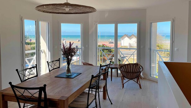 A dining area with a wooden table and chairs, a vase with flowers, and large windows offering a view of the beach and ocean. - Entire Seaside House, 160m² – 3 Bedrooms, Sleeps 6 (Ouistreham)