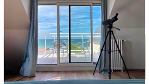 A room with a large sliding glass door leading to a balcony with a view of the sea. There is a telescope on a tripod on the balcony. - Entire Seaside House, 160m² – 3 Bedrooms, Sleeps 6 (Ouistreham)