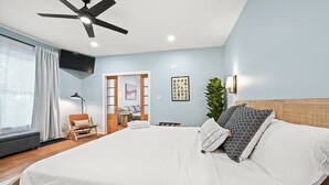 Ein Schlafzimmer mit Bett, Deckenventilator, Fenster mit Vorhängen, Sessel, Nachttisch und Wandlampe.