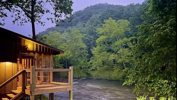 A wooden cabin with a deck overlooking a river, surrounded by lush greenery and hills.
