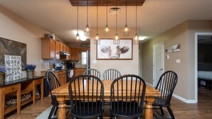A dining area with a wooden table and chairs, a kitchen in the background, and pendant lights hanging from the ceiling. - On River/Mt. LeConte Views/DwTn (Pigeon Forge)