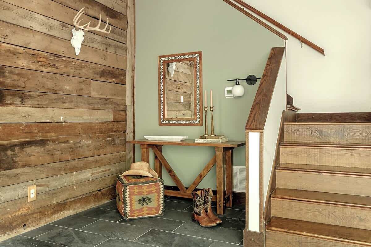 A rustic interior with a wooden wall, a mirror, a table with candles, a woven basket, and a pair of boots.
