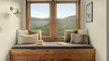 A wooden bench with a checkered cushion, a hat, and an open book, positioned in front of a large window with a view of a forested landscape.
