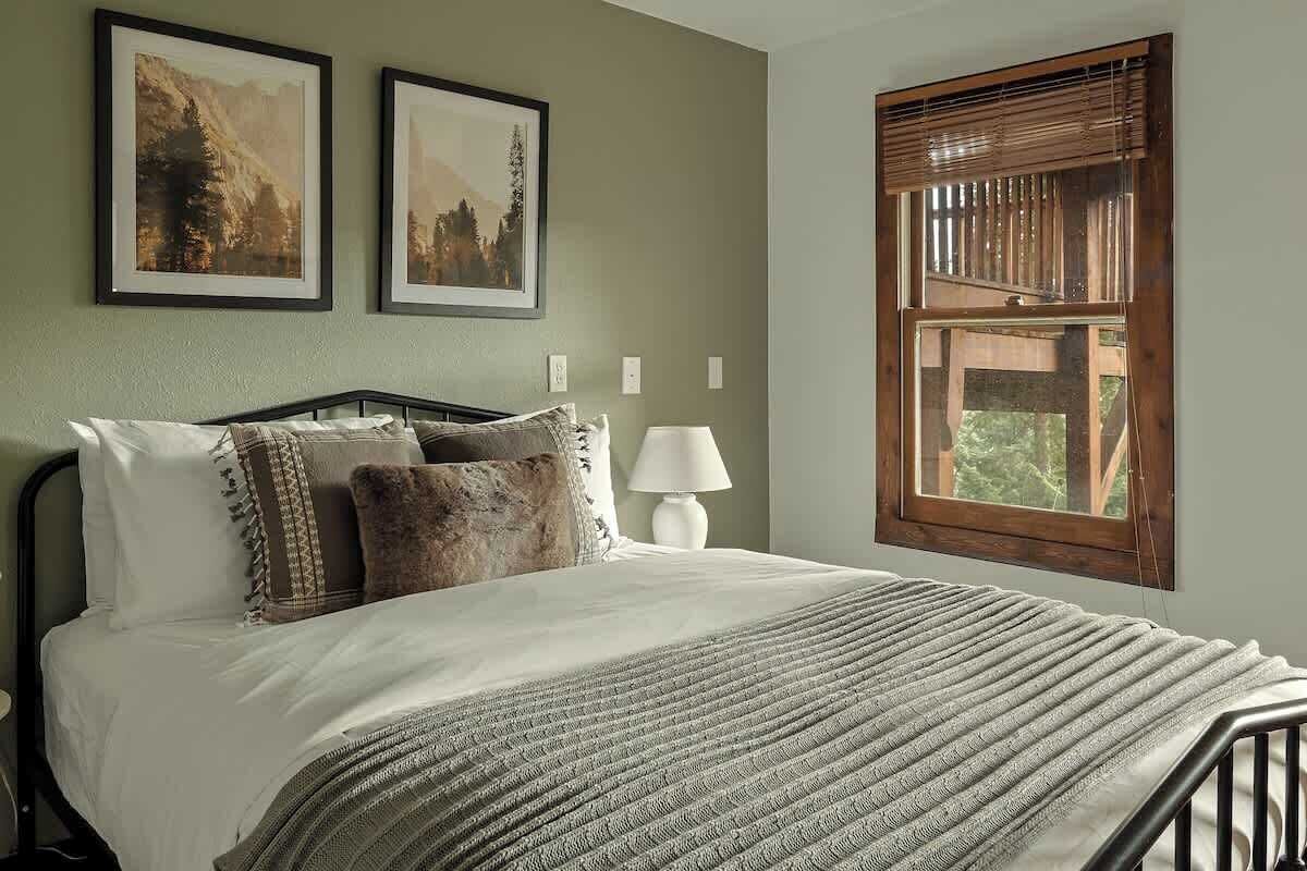 A bedroom with a bed, two framed pictures, a window with blinds, and a bedside lamp.