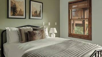 A bedroom with a bed, two framed pictures, a window with blinds, and a bedside lamp.