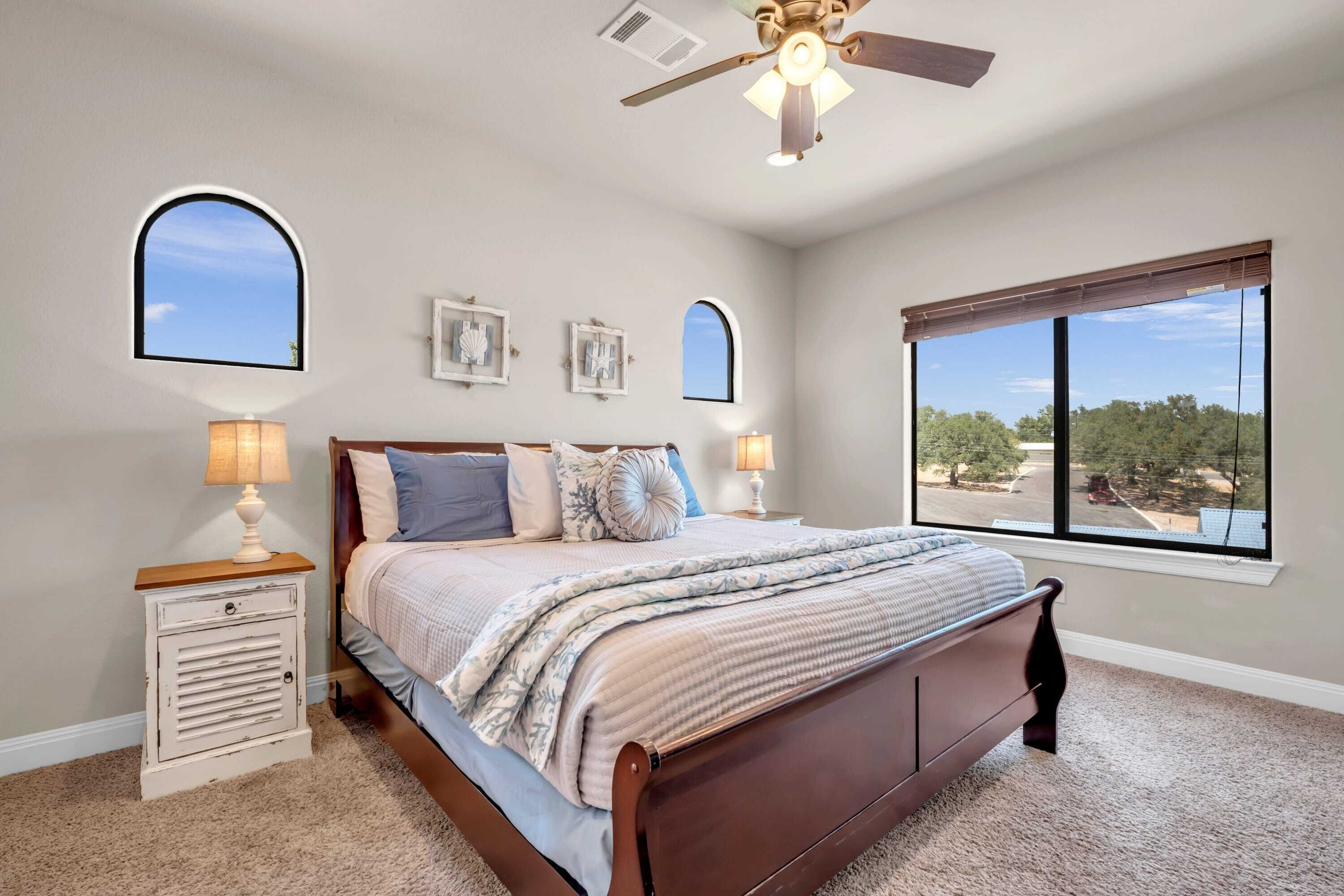 A bedroom with a bed, two bedside lamps, a ceiling fan, and a large window with a view.