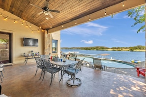 Une terrasse extérieure avec une piscine, une table entourée de chaises, un ventilateur de plafond et un téléviseur fixé au mur.