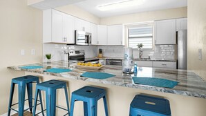 A modern kitchen with white cabinets, a marble countertop, and blue bar stools. - 🌊Fully Renovated | 500ft to Beach | 2 BR | Beach Within Reach🌊 (Miramar Beach)