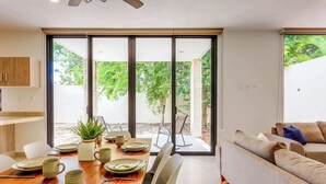 Uma área de refeições moderna com uma mesa de madeira posta para quatro pessoas, um sofá e vista para o verde através de grandes portas de vidro deslizantes.