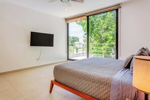 A bedroom with a bed, a television mounted on the wall, and a balcony with a view of trees.