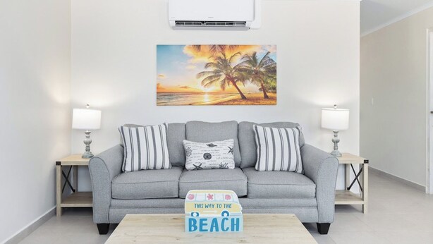Un salon avec un canapé gris, deux tables d’appoint avec des lampes, un tableau représentant une scène de plage et une table basse en bois sur laquelle se trouve une boîte décorée sur le thème de la plage.