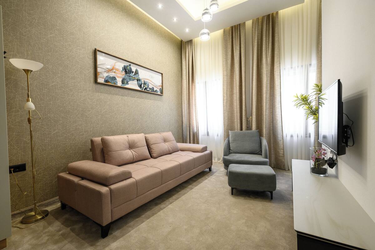 A modern living room with a beige sofa, a grey armchair, a floor lamp, a framed artwork, and a TV mounted on the wall.