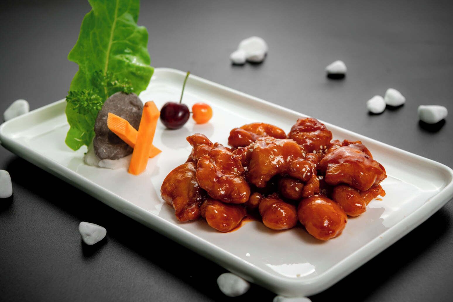 Une assiette rectangulaire blanche sur laquelle on a disposé un plat de poulet et de légumes glacés, le tout agrémenté d’une feuille et d’une cerise, le tout posé sur une surface sombre.