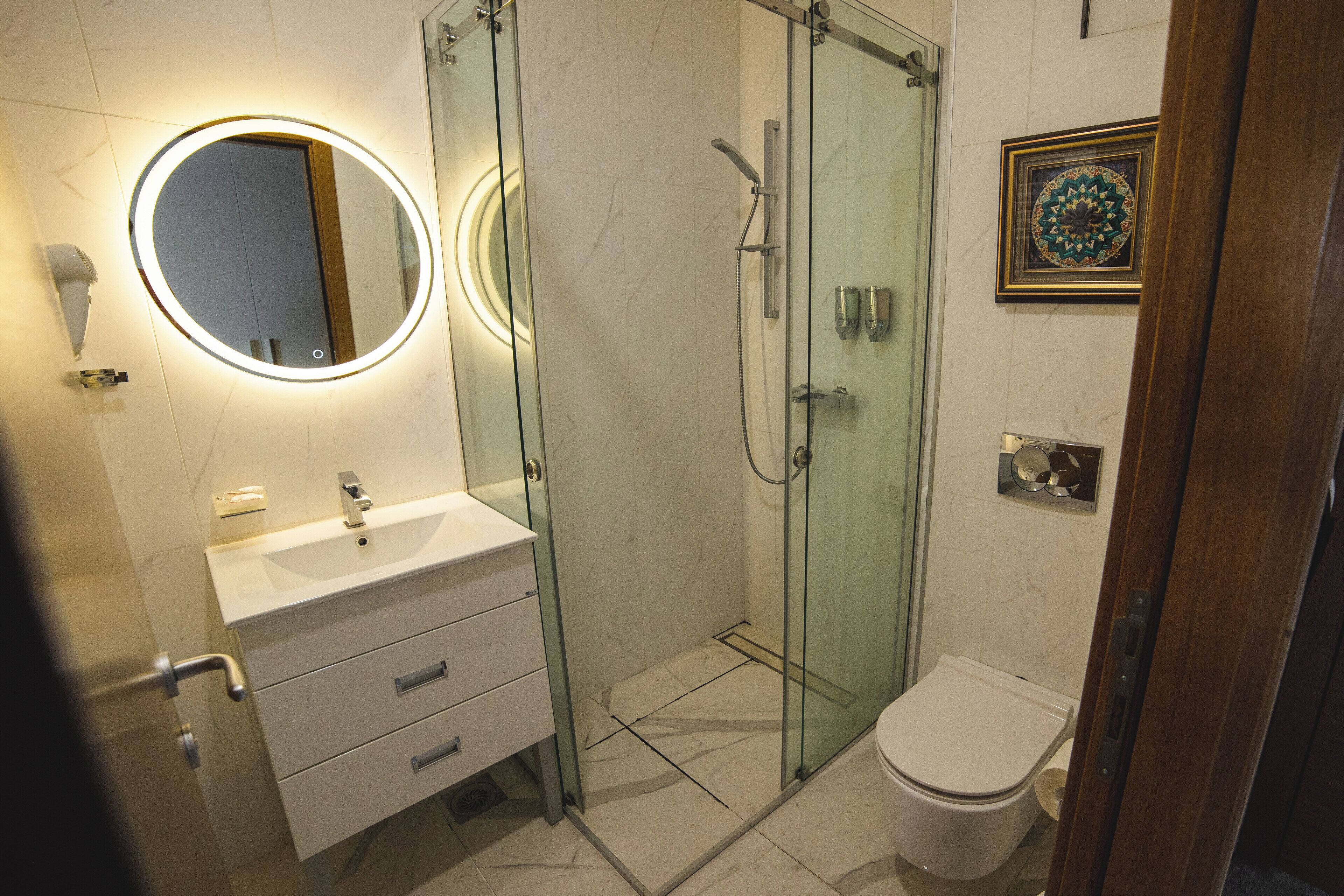 Un baño moderno con ducha de vidrio, un espejo redondo con iluminación, un lavamanos blanco y un inodoro.