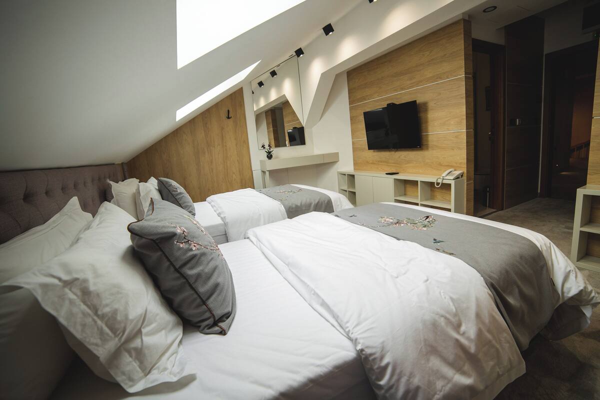 A modern bedroom with a sloped ceiling, a large bed with white bedding, a bedside table with a phone, and a TV mounted on the wall.