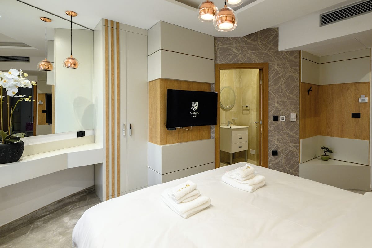 A modern hotel room with a large bed, a flat-screen TV, a wooden vanity, and a large mirror.