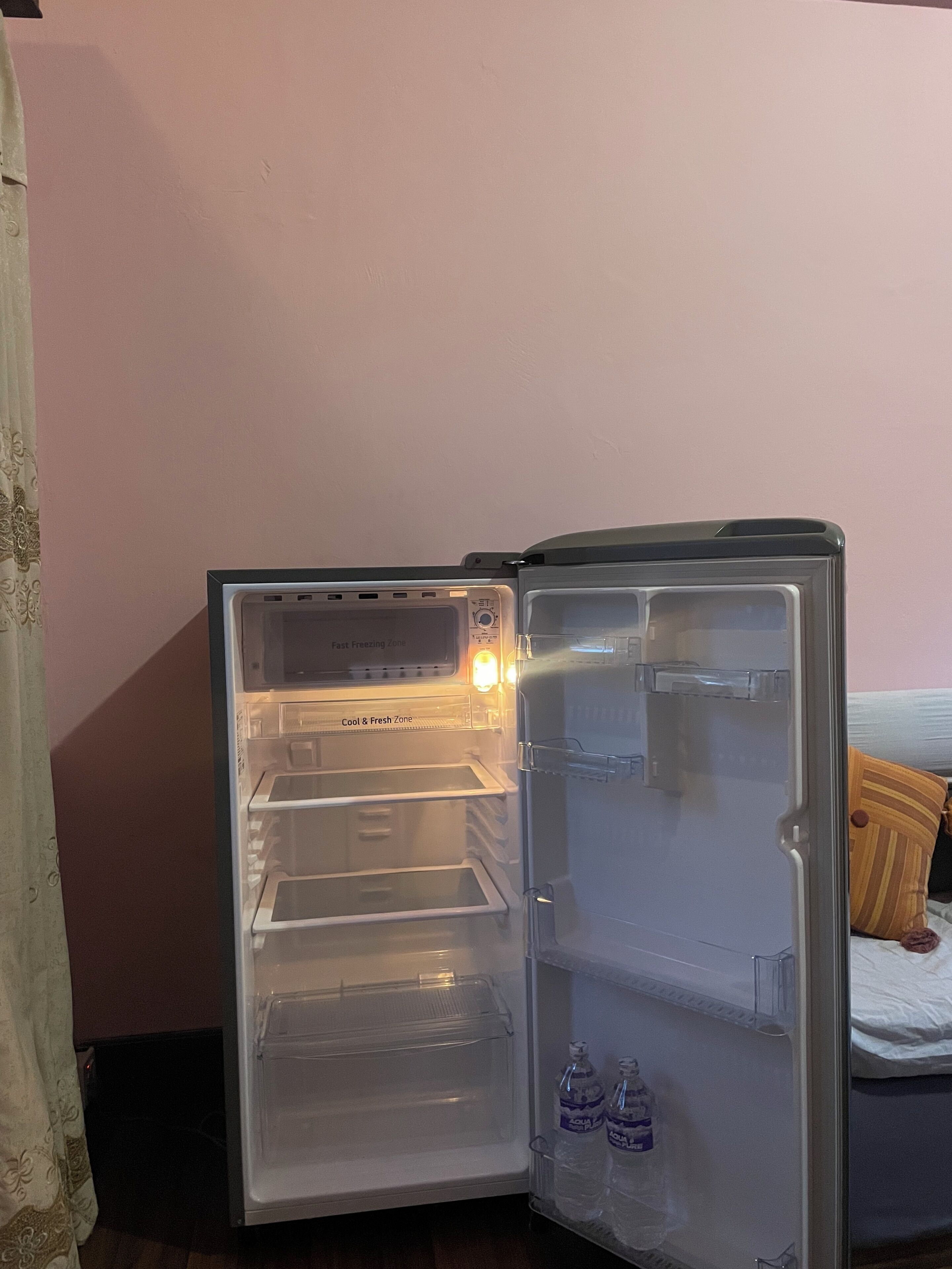 An open refrigerator with a "Cool & Fresh Zone" label, two water bottles, and a bed with a striped pillow in the background.