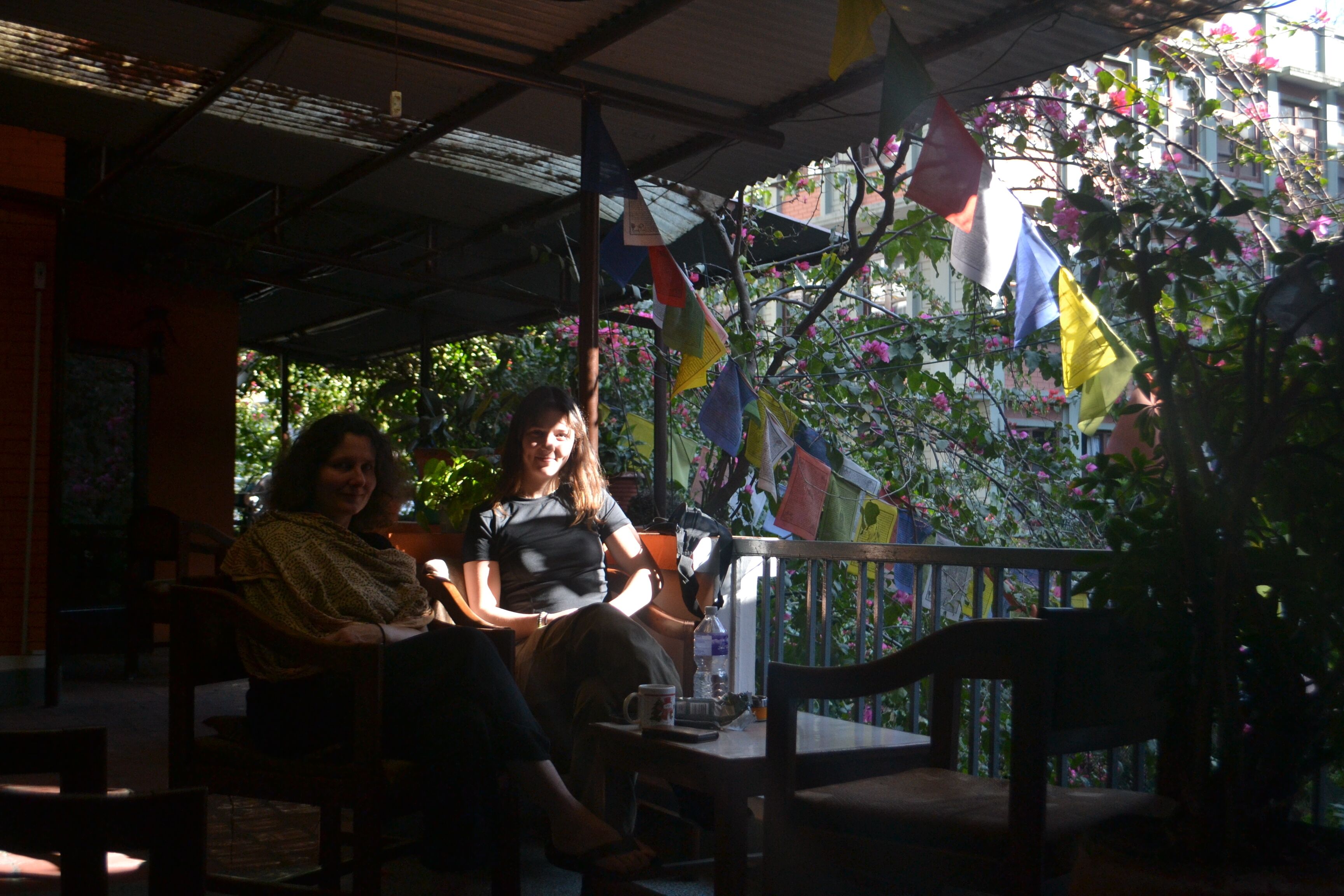 A covered outdoor seating area with chairs and tables, surrounded by greenery and colorful flags.