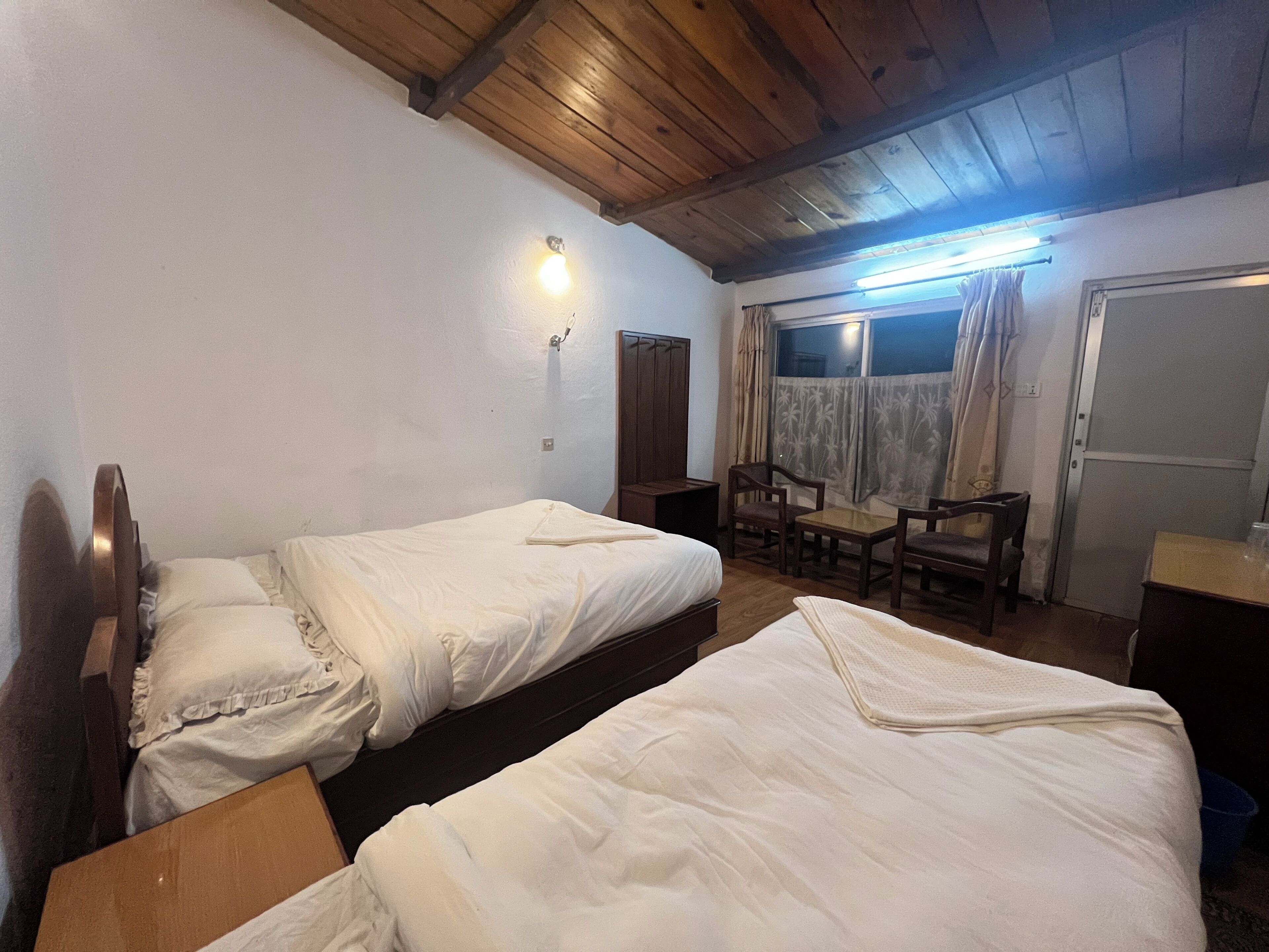 A room with two beds, a wooden ceiling, a window with curtains, and a small table with chairs.