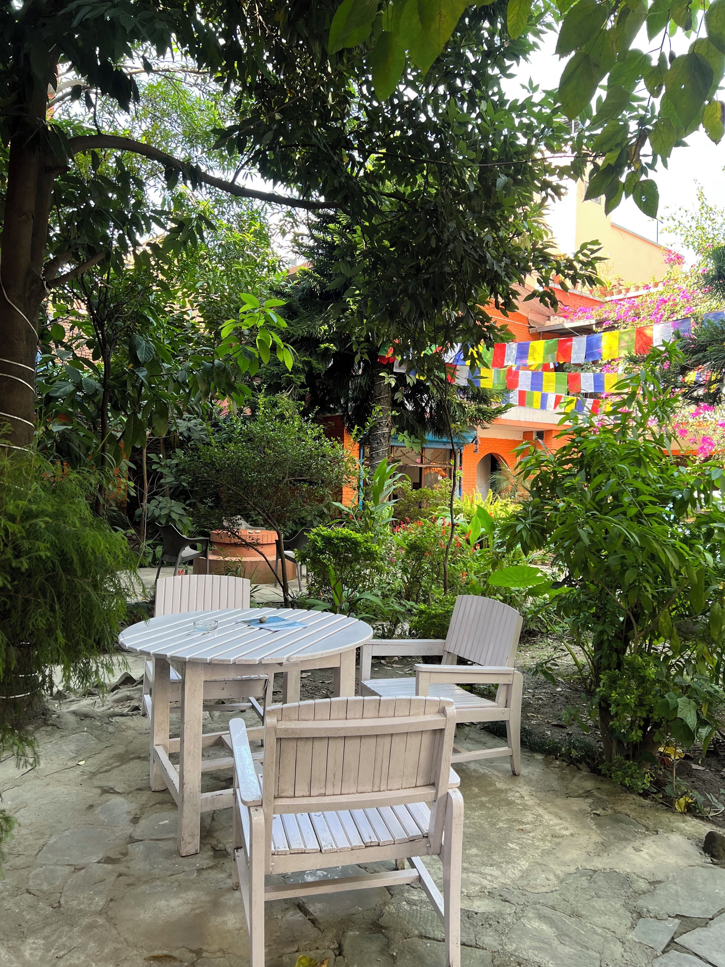 Un área de estar al aire libre con una mesa redonda y dos sillas, rodeada de abundante follaje y un edificio con decoraciones coloridas.