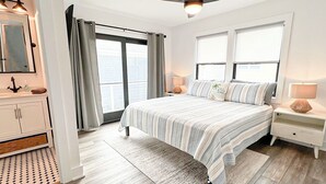 Un dormitorio moderno con una cama grande, ventilador de techo, mesita de noche y baño con tocador y espejo.