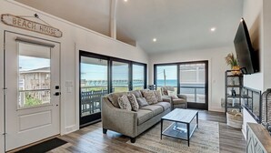 Amplia sala de estar con un sofá grande, una mesa de centro y un televisor montado en la pared. Se observa una puerta con la etiqueta "Beach House" y ventanas grandes que ofrecen vista a la playa y al océano.