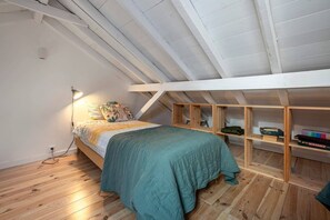 Un dormitorio con techo inclinado, piso de madera, cama con cubrecama verde y estante con libros y un juego de Scrabble.